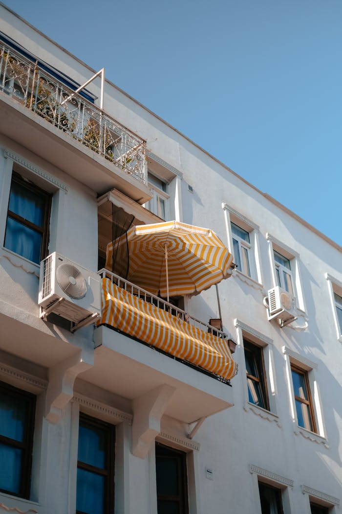 Low-angle view of a white building with a classic balcony featuring a yellow striped umbrella.
